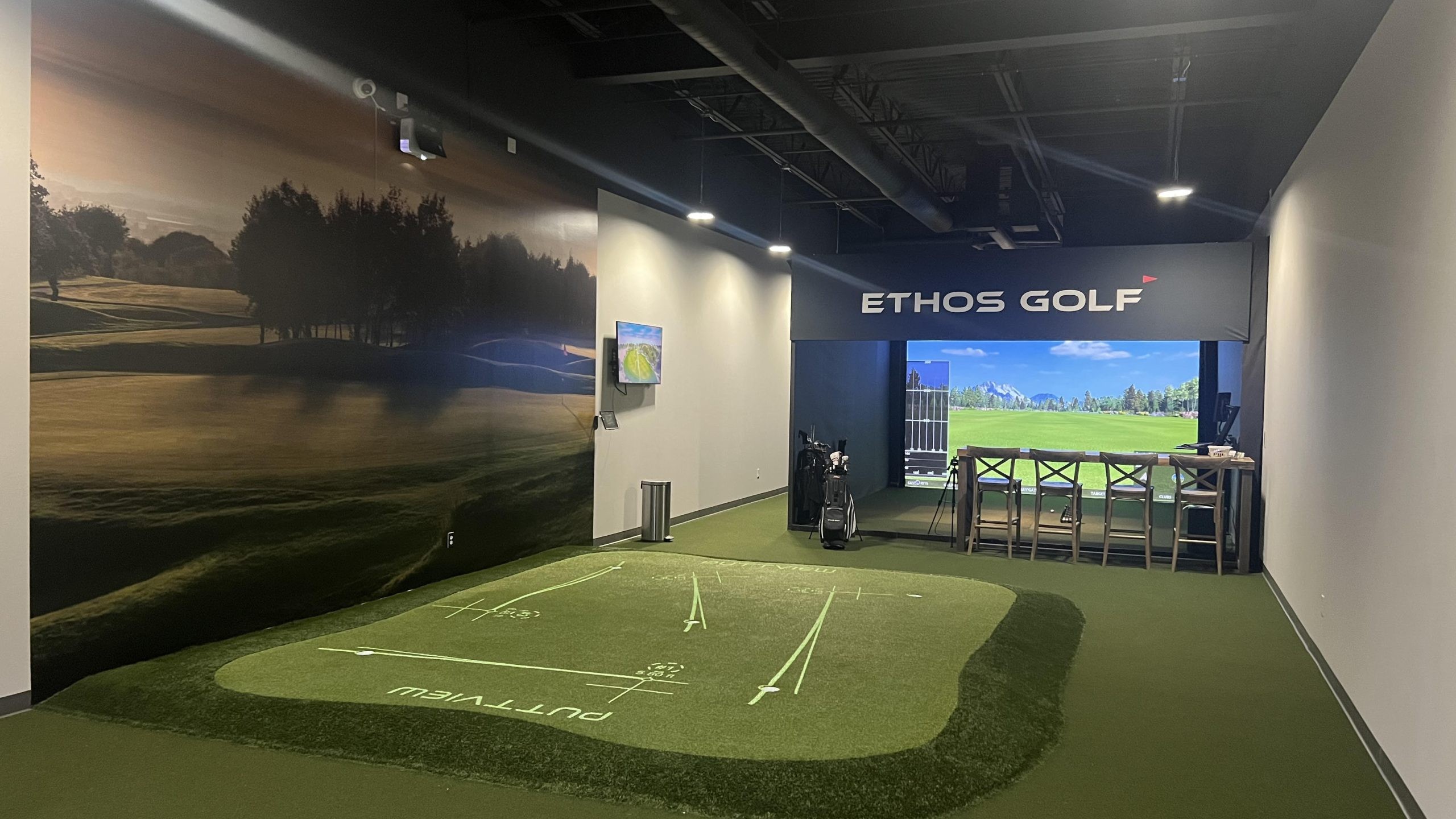 A sleek indoor golf simulation room with a putting green area, a large screen showing a virtual golf course, and golf clubs and bags in the middle. The wall on the left features a mural of an outdoor golf course, recreating the vibe of Allen Texas Golf. 